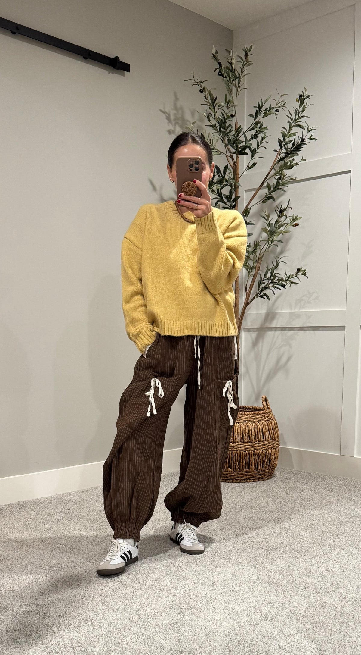 Person wearing a yellow sweater and brown pants taking a mirror selfie in a room with a plant and woven basket.