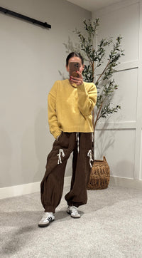Person wearing a yellow sweater and brown pants taking a mirror selfie in a room with a plant and woven basket.
