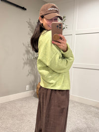 Person taking a mirror selfie wearing a light green sweater and brown pants in a room with white walls and a wooden floor.